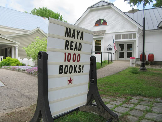 Outdoor sign in front of a white building reads "MAYA READ 1000 BOOKS!" with the number in red letters and a red star below the text.