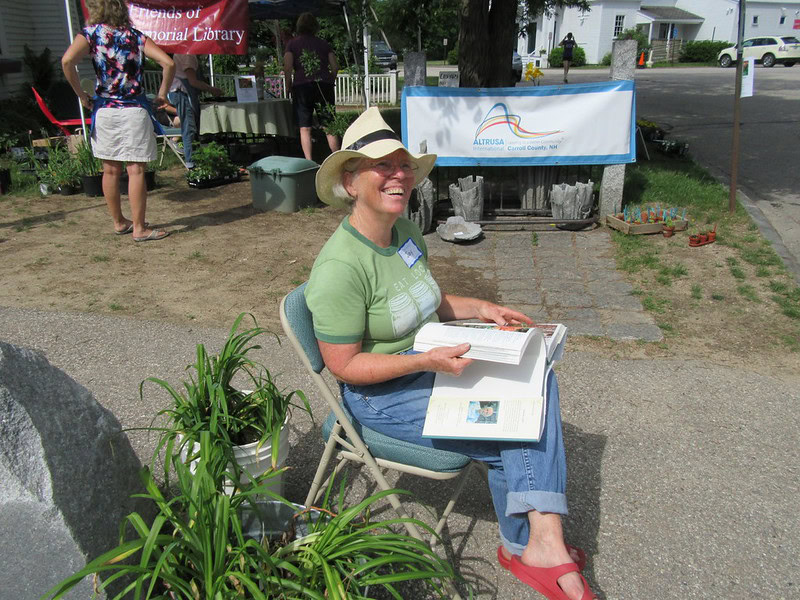 Photo of Jay Rancourt, sitting on a chair in front of the Cook Memorial Library holding an open book in her lap. Taken at the annual plant sale held in 2016.