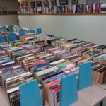 Rows of assorted books are organized on tables, separated by blue bookends, in a well-lit room lined with more bookshelves.