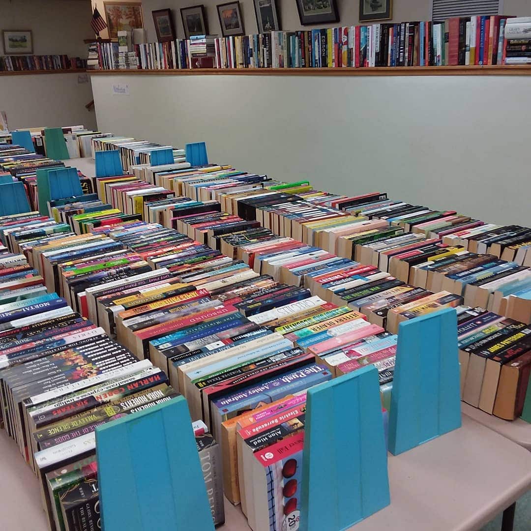 Rows of assorted books are organized on tables, separated by blue bookends, in a well-lit room lined with more bookshelves.