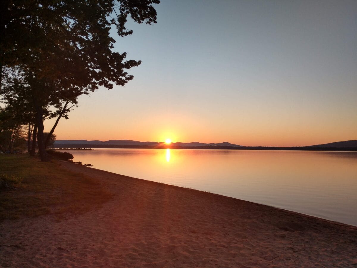 Photo of a sandy beach by a lake with the sun rising over mountains in the distance