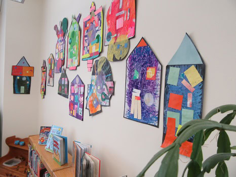 Colorful paper house collages are displayed on a wall above a bookshelf filled with children's books, with part of a green plant visible in the corner.