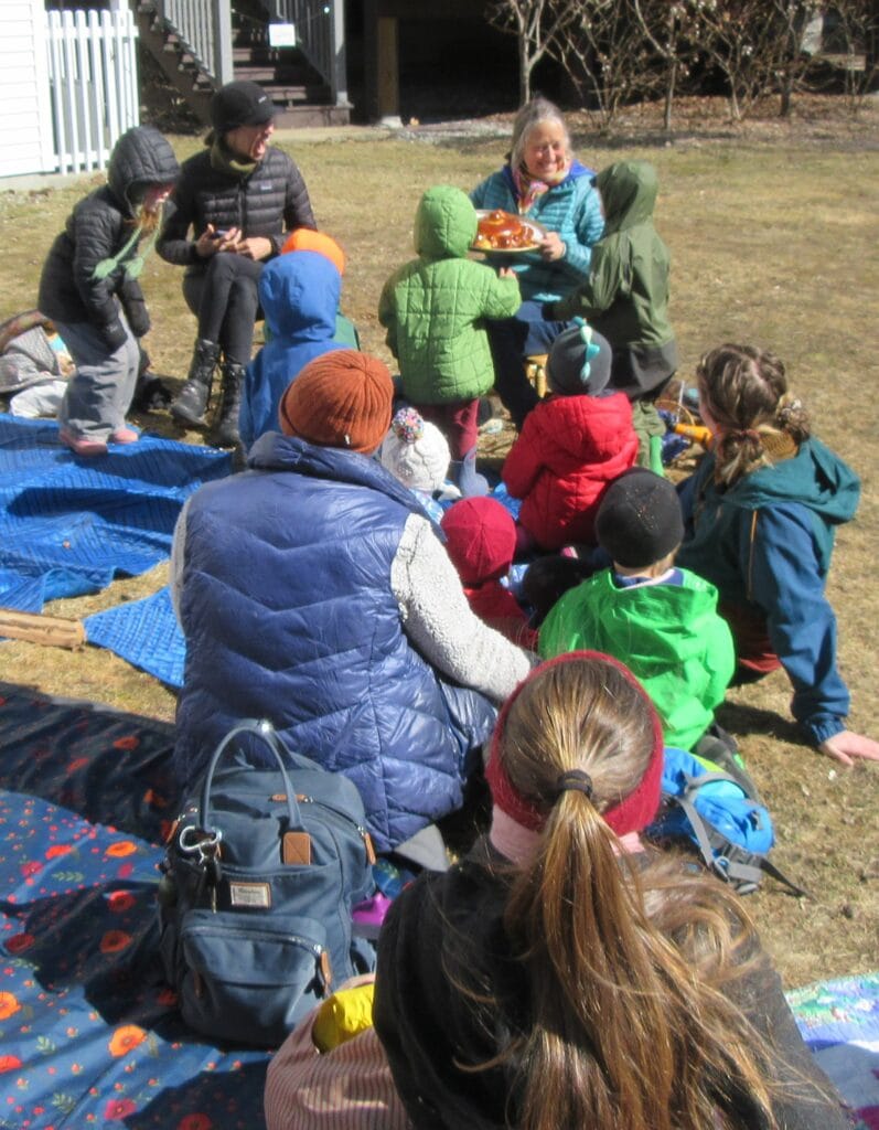 A group of adults and children sit on blankets outside on a sunny day, gathered around a woman holding a tray of food.