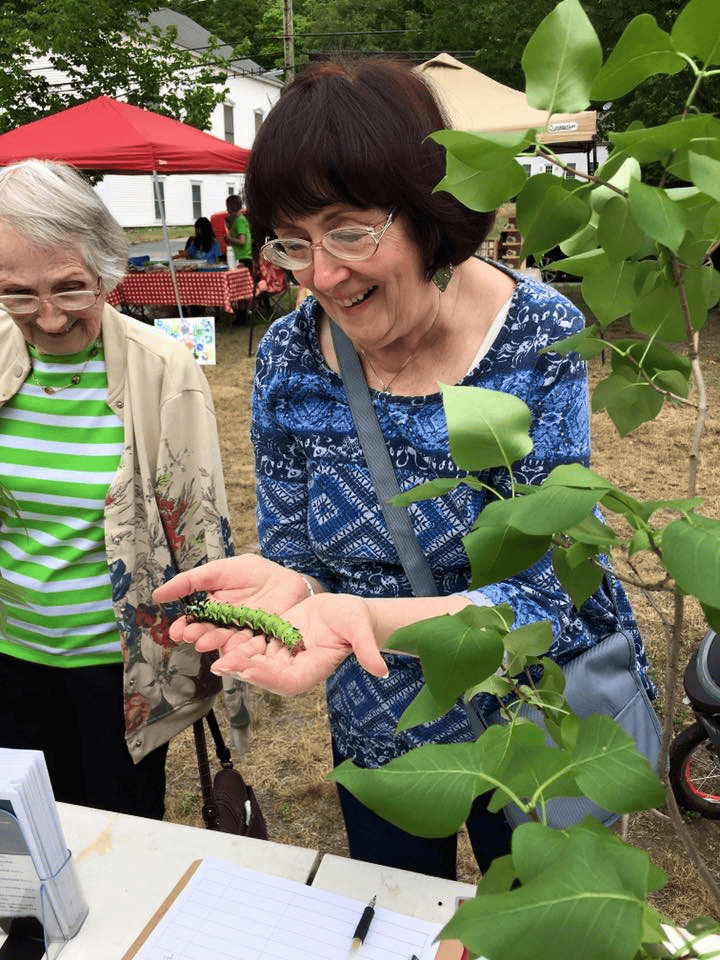 Two women stand outdoors at a market or fair, one smiling while holding a large green caterpillar on her hand near a leafy plant.